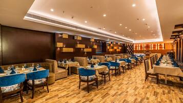 A spacious dining area with multiple tables and chairs, a high ceiling with recessed lighting, and a wooden floor.