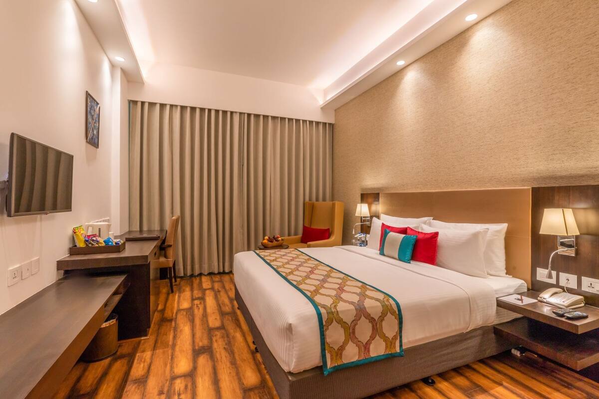 Quarto de hotel com uma cama grande, uma escrivaninha, uma cadeira, uma televisão e uma janela com cortinas.