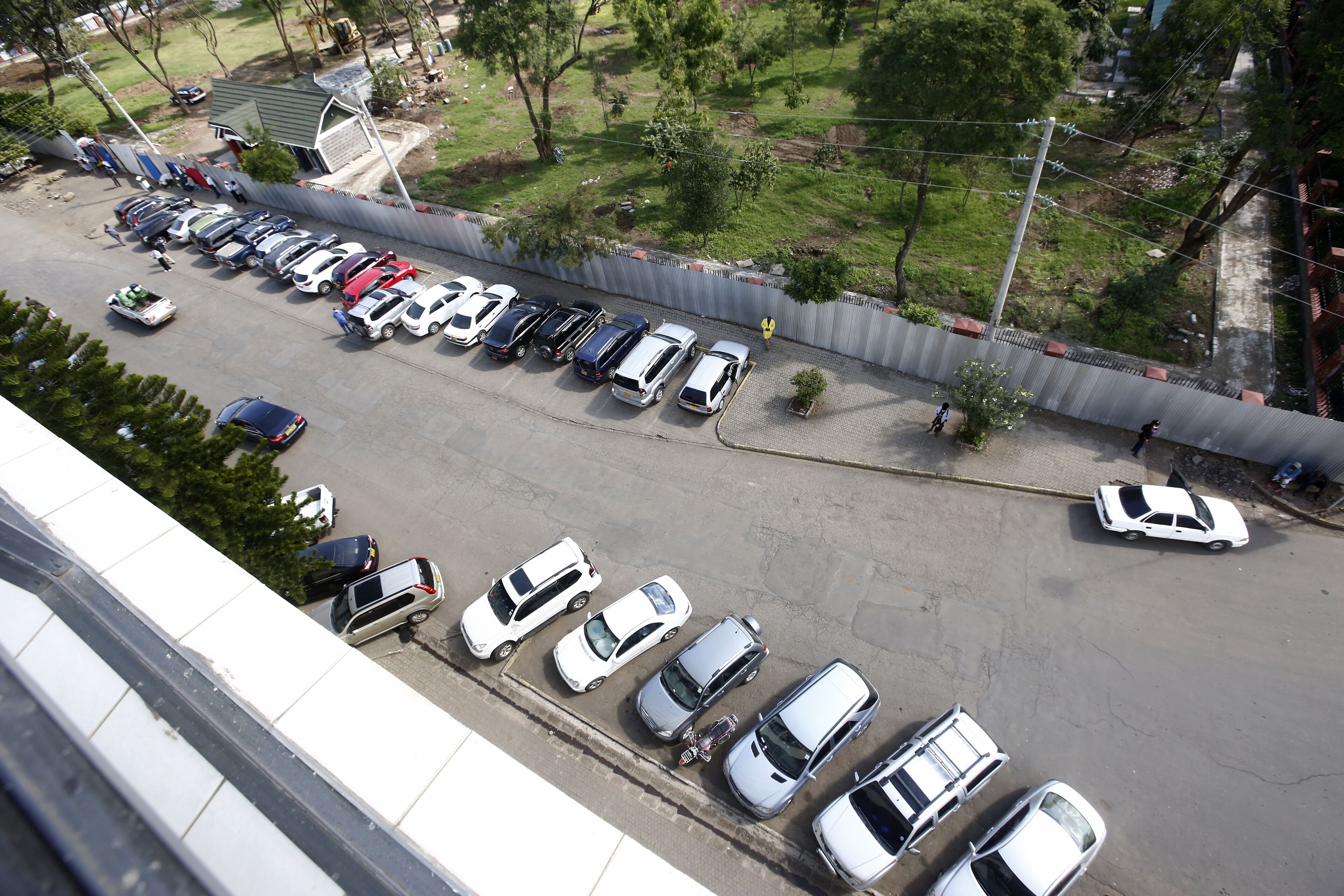 Vista aérea de un estacionamiento con varios coches aparcados en filas, una zona verde con árboles y una valla delimitando el terreno.