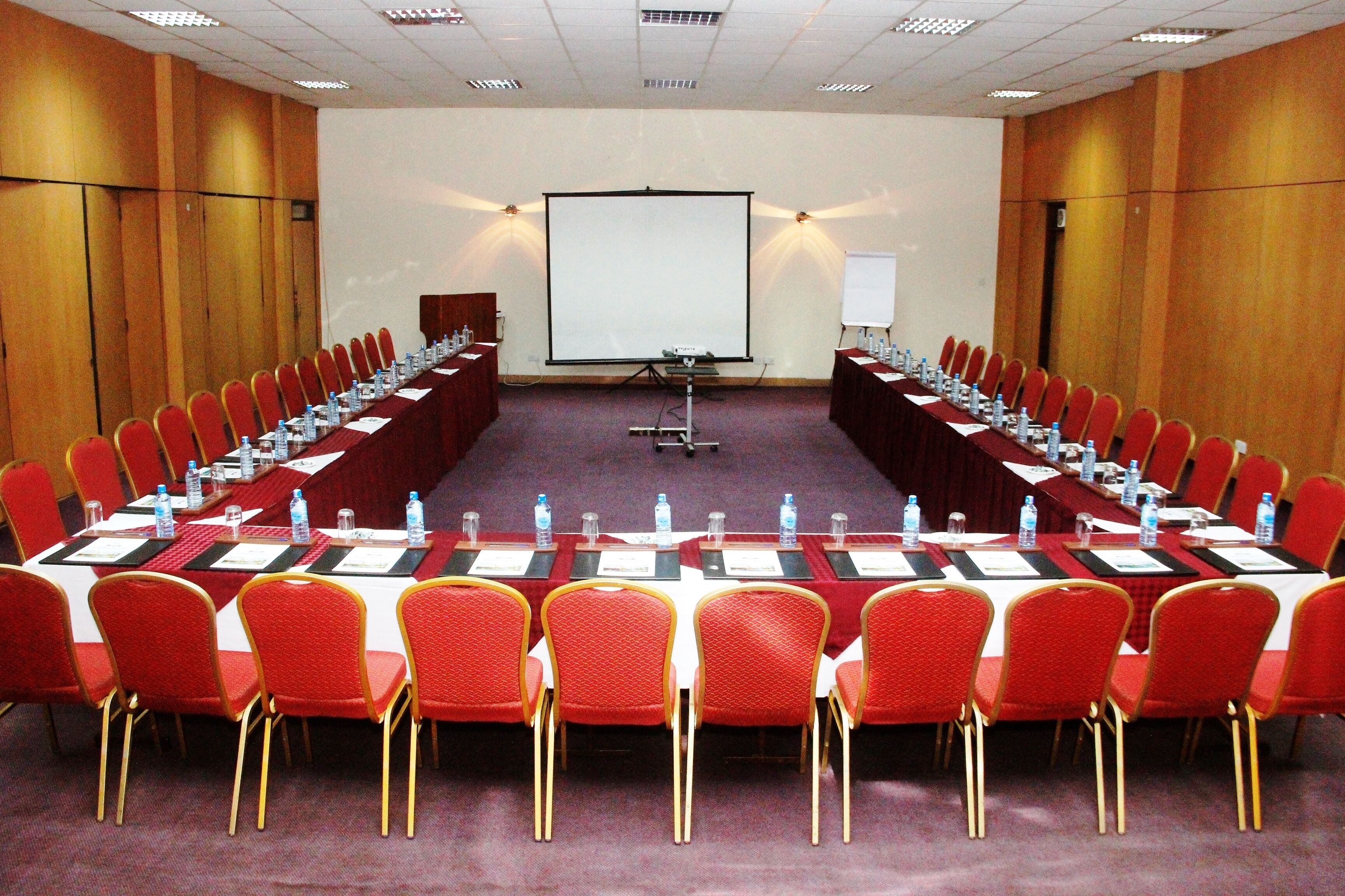 Un salón de conferencias con una larga mesa, sillas rojas, botellas de agua y una pantalla de proyector.