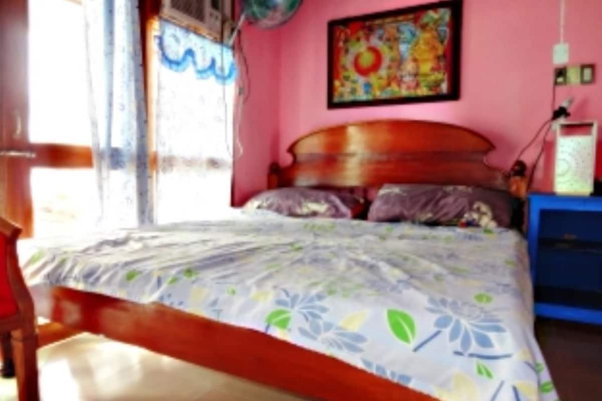 A bedroom with a wooden bed frame, floral bedding, a colorful painting on the wall, and a blue nightstand with a lamp.