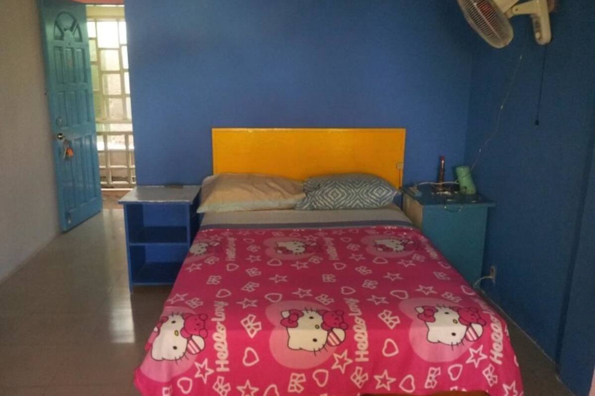 A bedroom with a bed covered in a Hello Kitty themed bedspread, a bedside table with a lamp, and a wall-mounted fan.