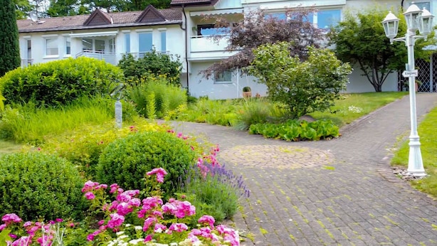 A residential building with a paved pathway, lush greenery, and flowering plants. - Hotel Haus Am Steinberg (Bad Driburg)