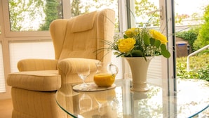 A room with a beige armchair, a glass table with a pitcher of orange juice, and a vase with yellow flowers. - Hotel Haus Am Steinberg (Bad Driburg)