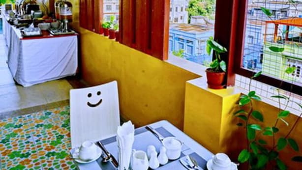 A dining area with a table set for two, a window with red curtains, and a view of buildings. - Hotel Aurora (Mandalay)
