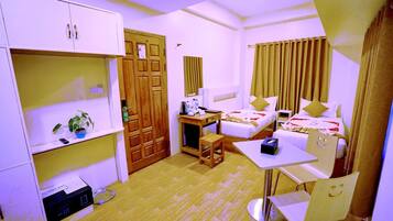 A compact hotel room with a single bed, a desk with a chair, a small table, a wardrobe, and a shelf with a plant.