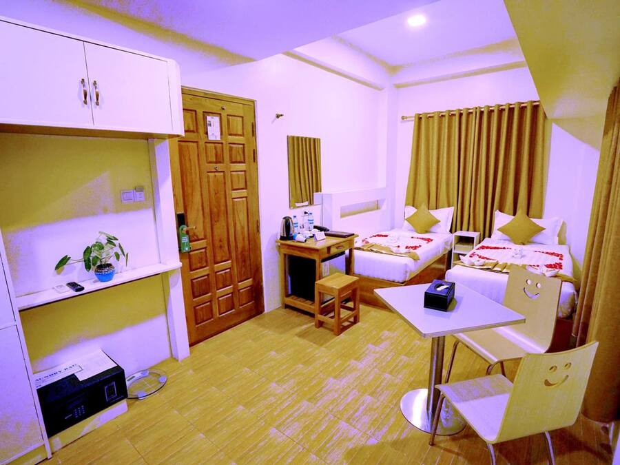 A compact hotel room with a single bed, a desk with a chair, a small table, a wardrobe, and a shelf with a plant.