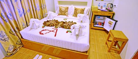 A hotel room with a neatly made bed, a bedside table with a refrigerator, a wooden stool, and a large mirror on the wall.