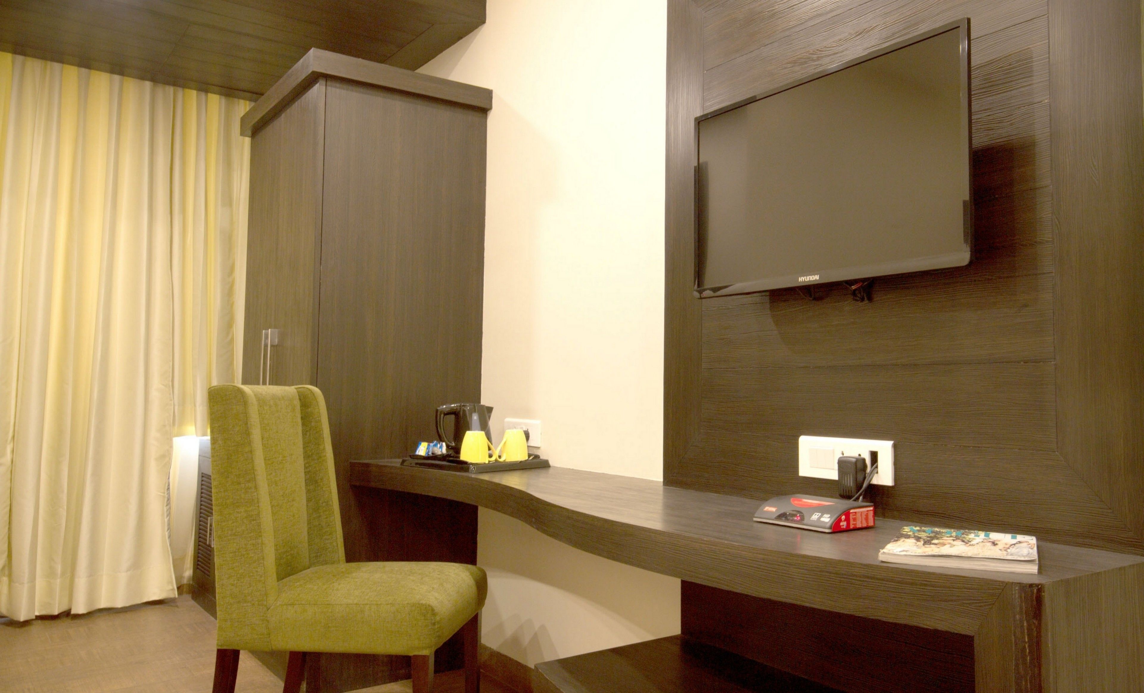 A hotel room with a wooden desk, a green chair, a flat-screen TV mounted on the wall, and a window with curtains.