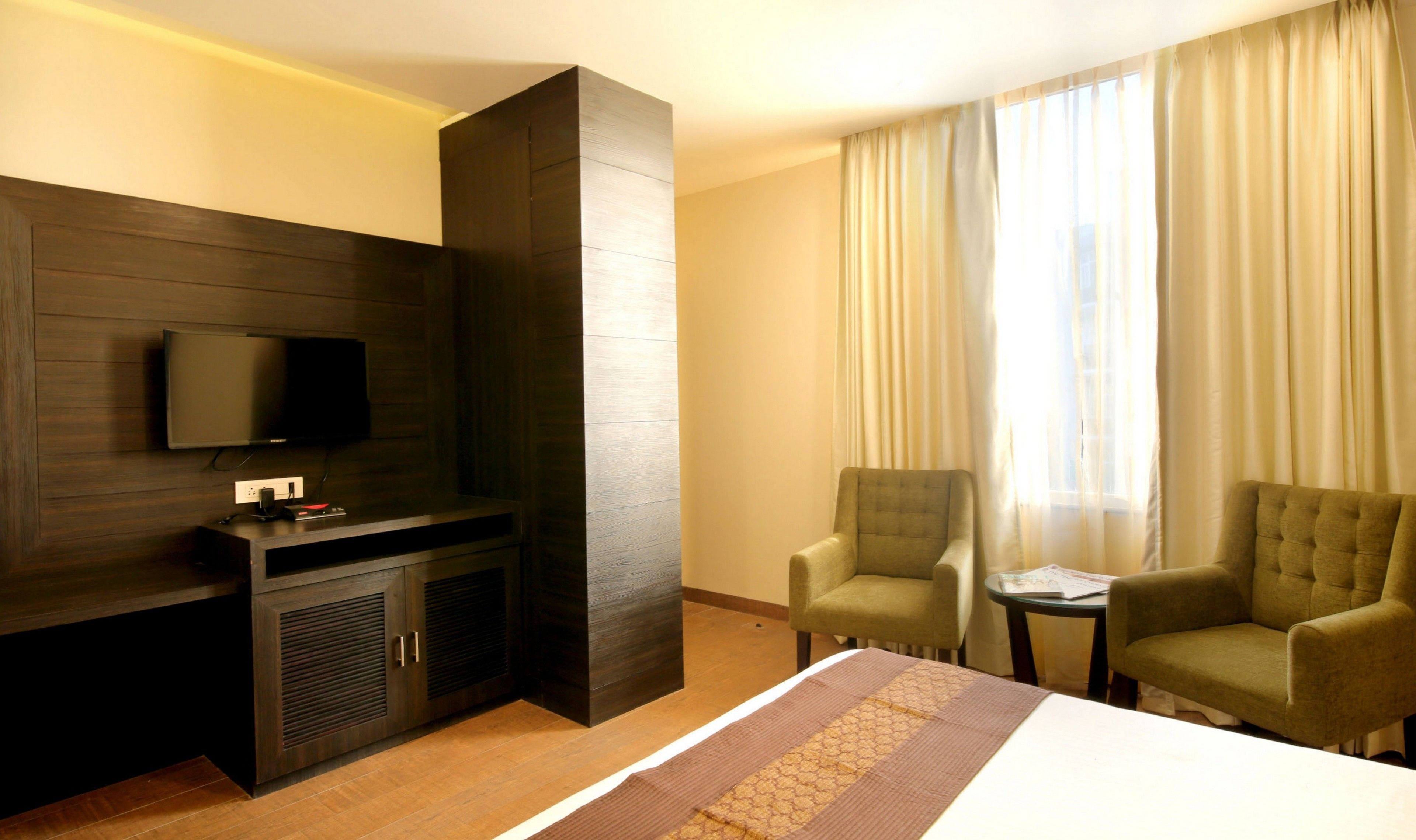 Hotelkamer met een flatscreen televisie aan een donkere houten wand, een bed met een geborduurde spreideken, twee fauteuils, een tafeltje met een tijdschrift en een groot raam met dunne gordijnen.