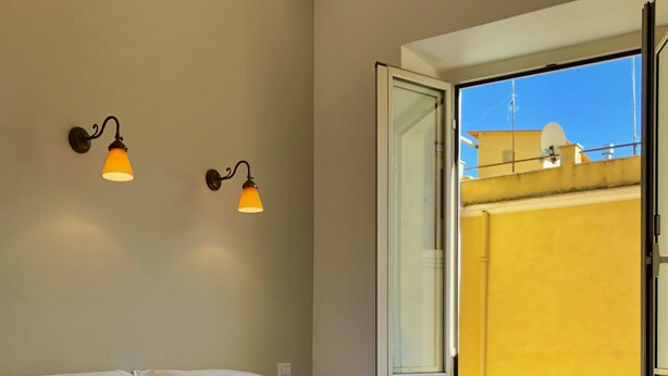 A neatly made bed with white linens and towels, a window with an open view, and two wall-mounted lamps. - Termini Station Little House (Rome)