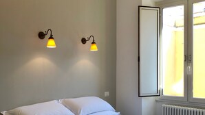 A neatly made bed with white linens, two wall-mounted lamps, a large mirror, and a window with curtains. - Termini Station Little House (Rome)