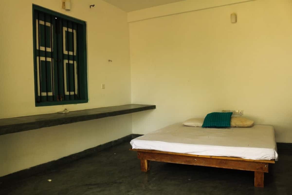 A single-bed room with a wooden frame, a window with metal bars, and a small shelf.