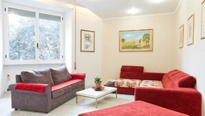 A living room with a grey sofa, a red sofa, and a red ottoman. There are two framed pictures on the wall and a window with blinds. - Flaminio & Popolo Large Apartment (Rome)