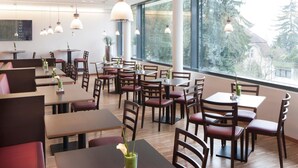 Een moderne eetruimte met meerdere tafels en stoelen, grote ramen met uitzicht op bomen en gebouwen, en hanglampen.