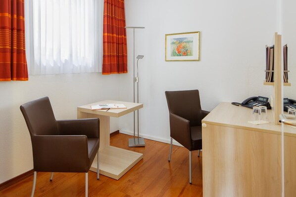A modern hotel room with two brown leather chairs, a small wooden table, a desk with a telephone and glass, and a floor lamp.