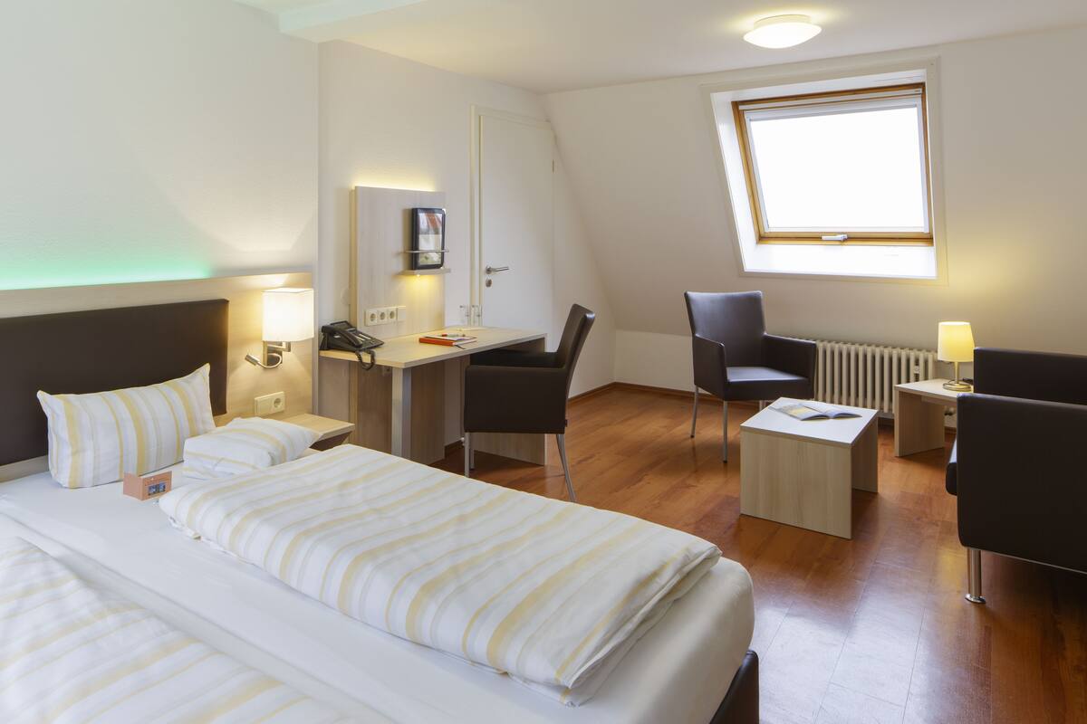 Habitación de hotel con una cama grande, un escritorio, dos sillas, una mesita y una ventana con luz natural.