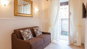Uma sala de estar com um sofá marrom, duas almofadas estampadas, um espelho com moldura dourada e vista através de uma porta com cortinas brancas.