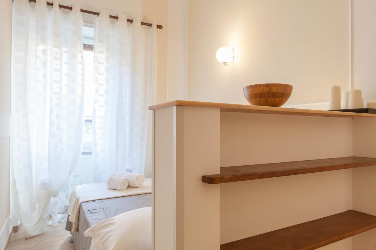 A hotel room with a bed, a wooden shelf with a bowl, and a wall-mounted light.