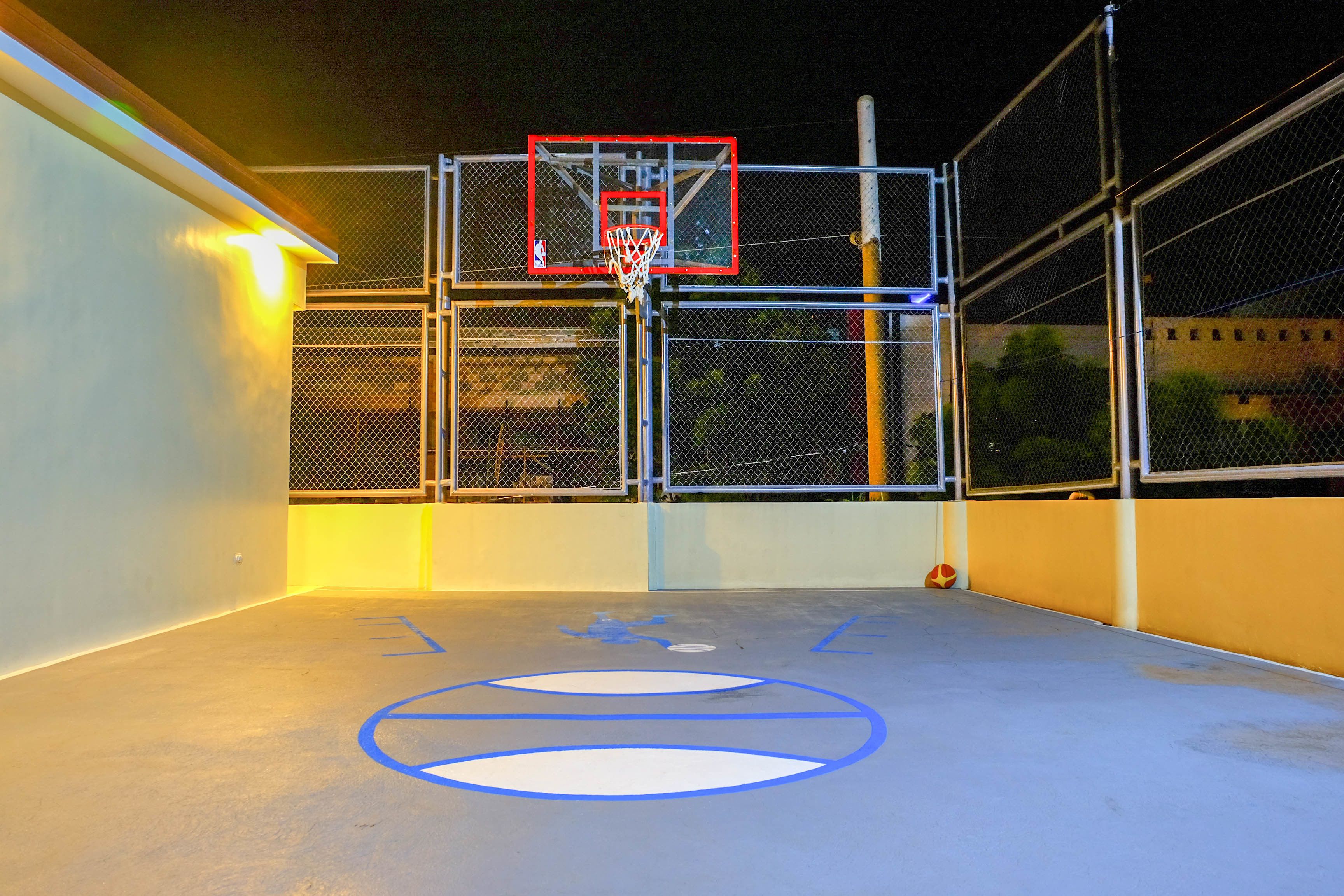 An outdoor basketball court with a hoop, surrounded by a chain-link fence, and marked with blue lines.