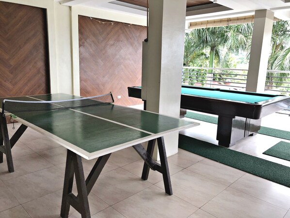 A billiards table and a pool table in a well-lit indoor area with tiled flooring and a view of greenery outside.