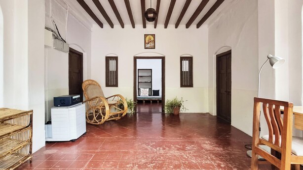 A room with a tiled floor, a bed with a patterned bedspread, a chair, a desk with a lamp, and a small cabinet. - Laika Heritage Stay by evaddo stays (Mysore)