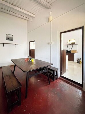A room with a wooden table, benches, a white ceiling, and a door leading to a kitchen area. - Laika Heritage Stay by evaddo stays (Mysore)