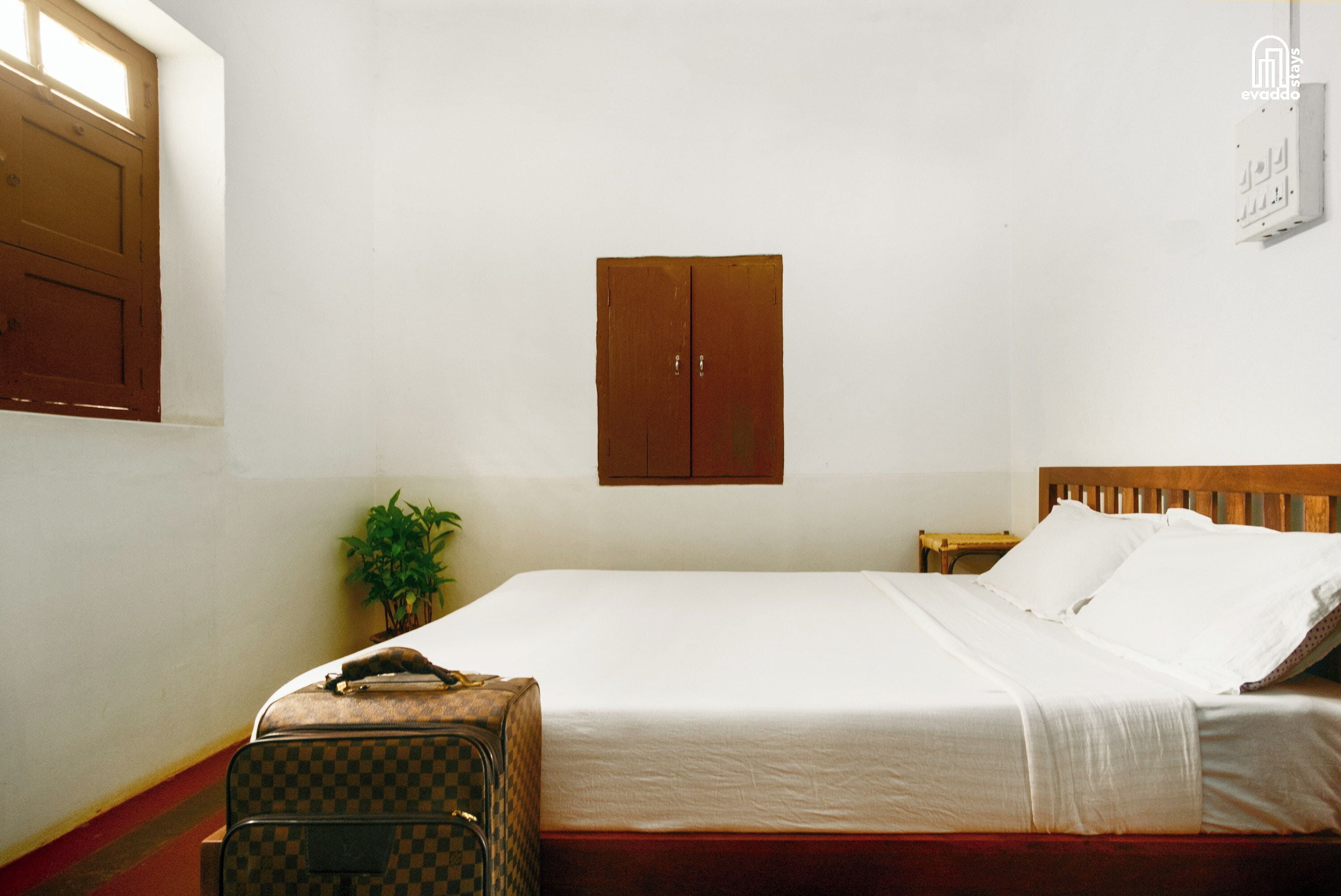 Una cama individual con ropa blanca, cabecera de madera, una mesita de noche pequeña, una maleta cuadrada marrón a cuadros, una planta en maceta y un armario de madera.