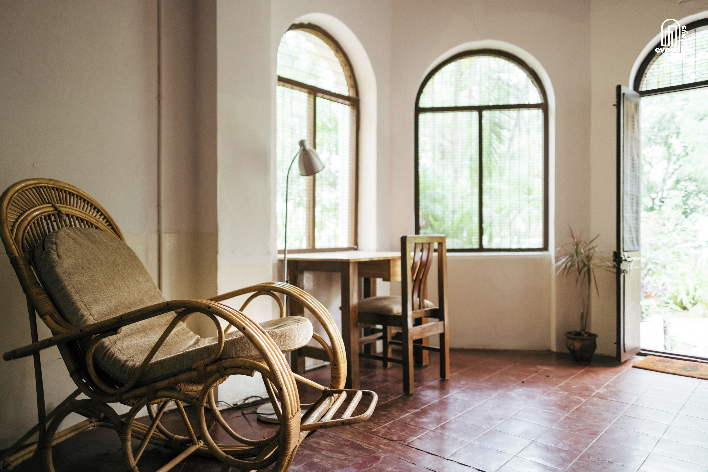 A room with a wicker chair, a wooden table, and two arched windows.