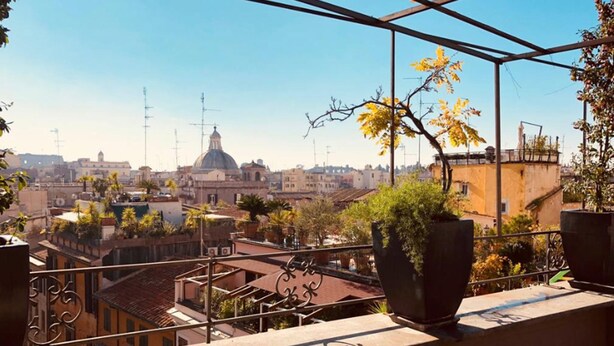 A balcony with potted plants, a wrought iron railing, and a view of a cityscape with buildings and a dome structure. - Il Covo (Rome)