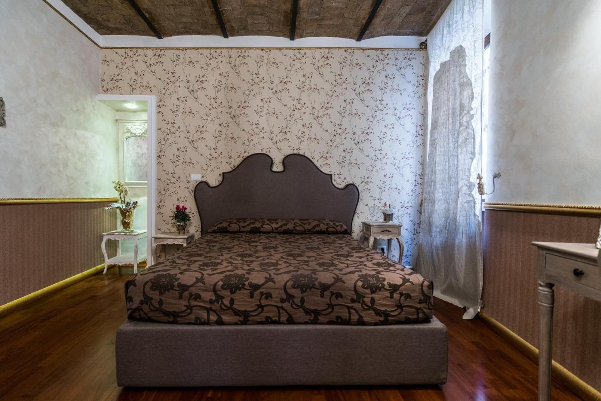 A bedroom with a large bed, floral wallpaper, a wooden floor, and a mirror.