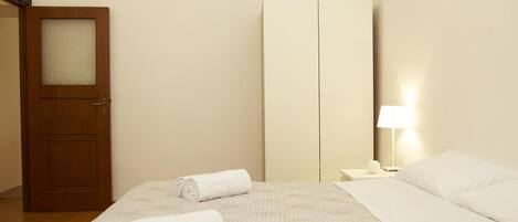 A hotel room with a bed, two rolled towels, a wooden door, a wardrobe, a nightstand with a lamp, and a wall-mounted light.