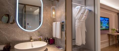 Un baño moderno con un lavamanos ovalado grande, un espejo con luz, zona de ducha y televisor.