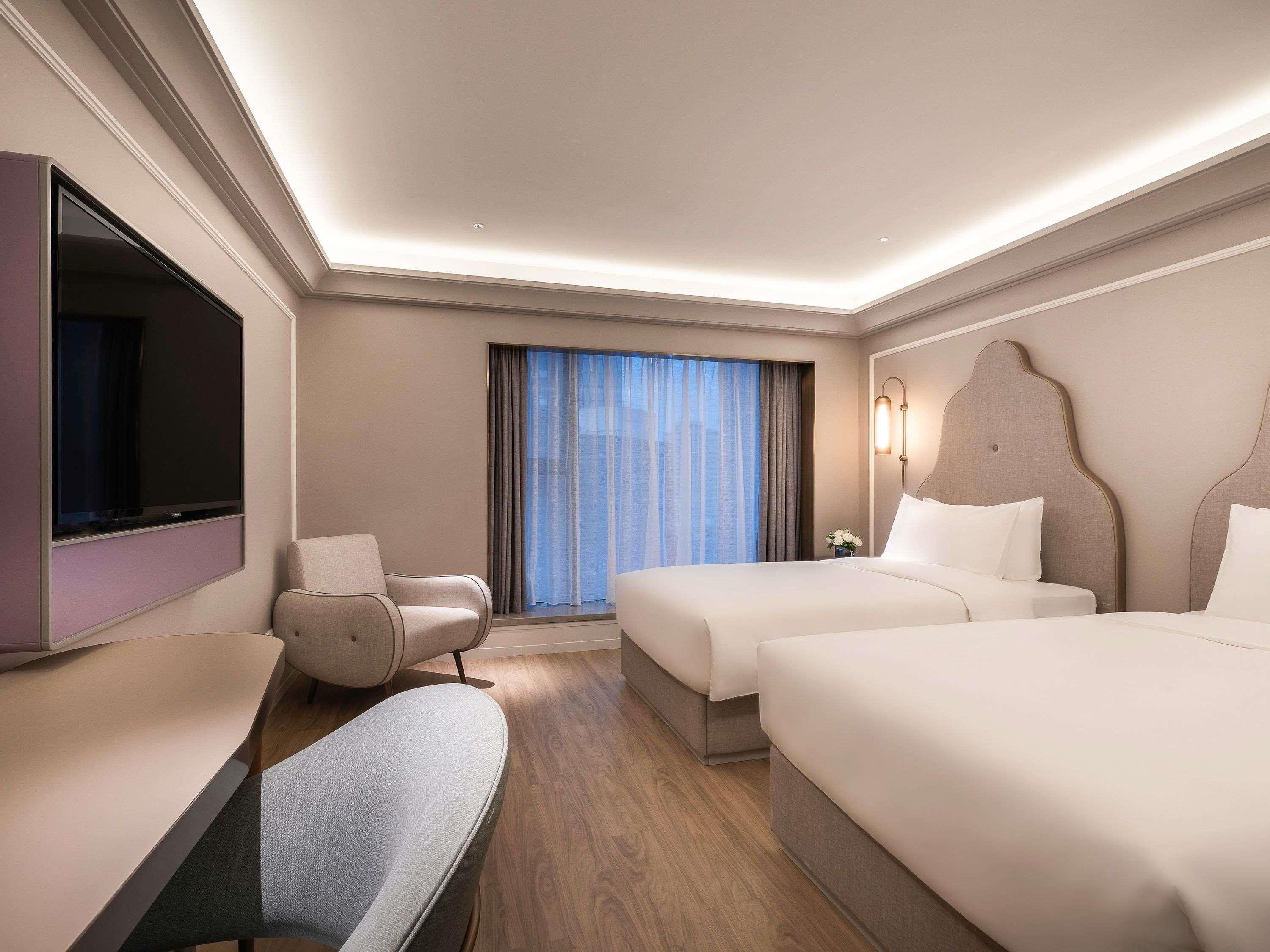 Ein Hotelzimmer mit Flachbildfernseher, einer Couch, zwei Betten, einem großen Fenster mit Vorhängen und einer Wandleuchte.