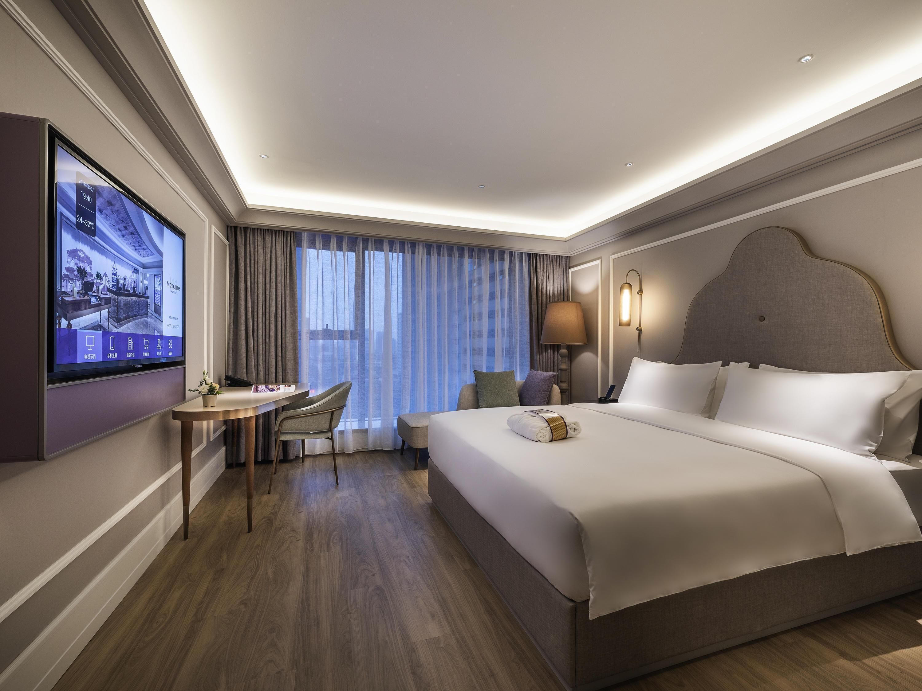 Habitación de hotel con una cama grande, un escritorio con silla, televisor montado en la pared y una ventana con cortinas.