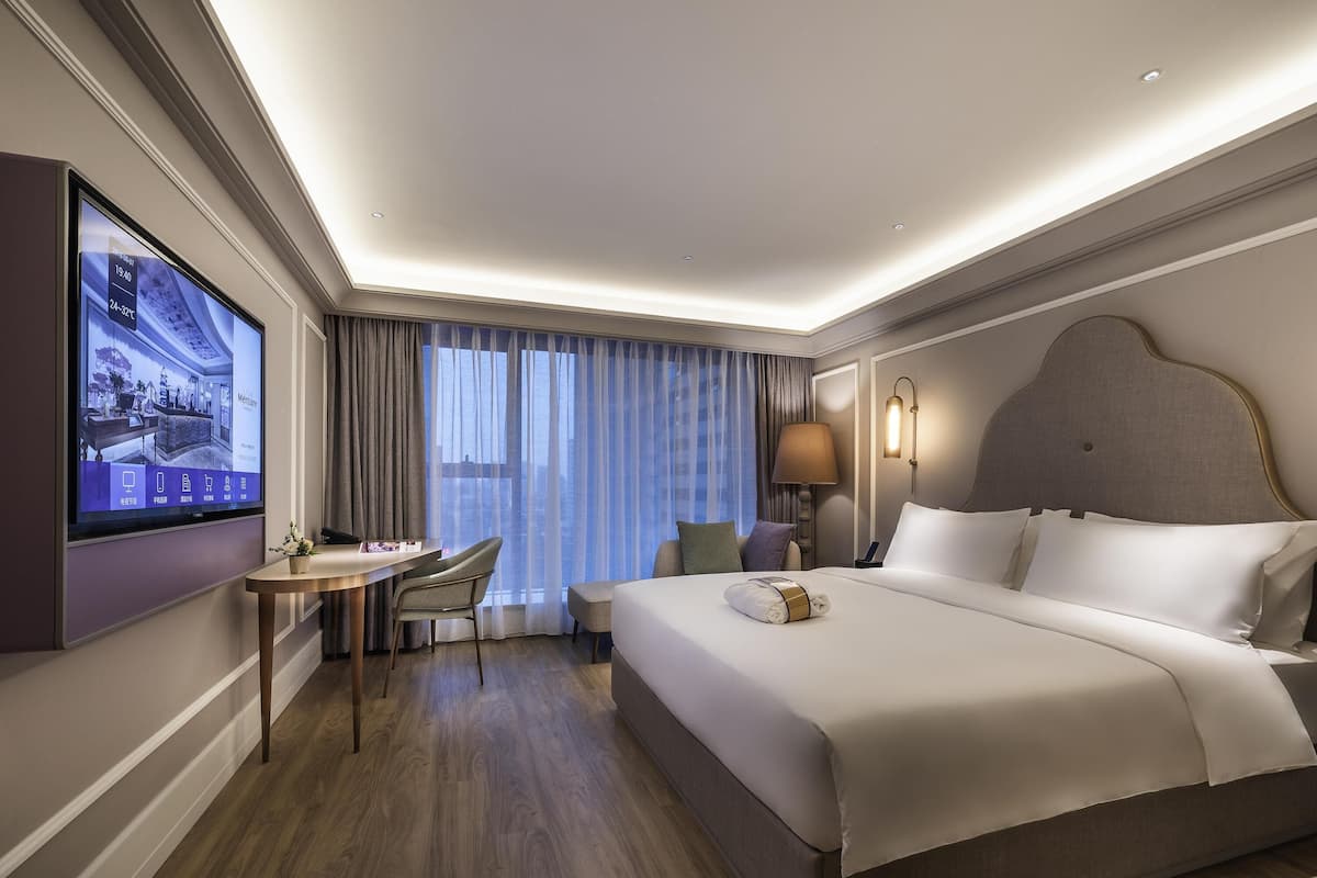 Habitación de hotel con una cama grande, un escritorio con silla, televisor montado en la pared y una ventana con cortinas.