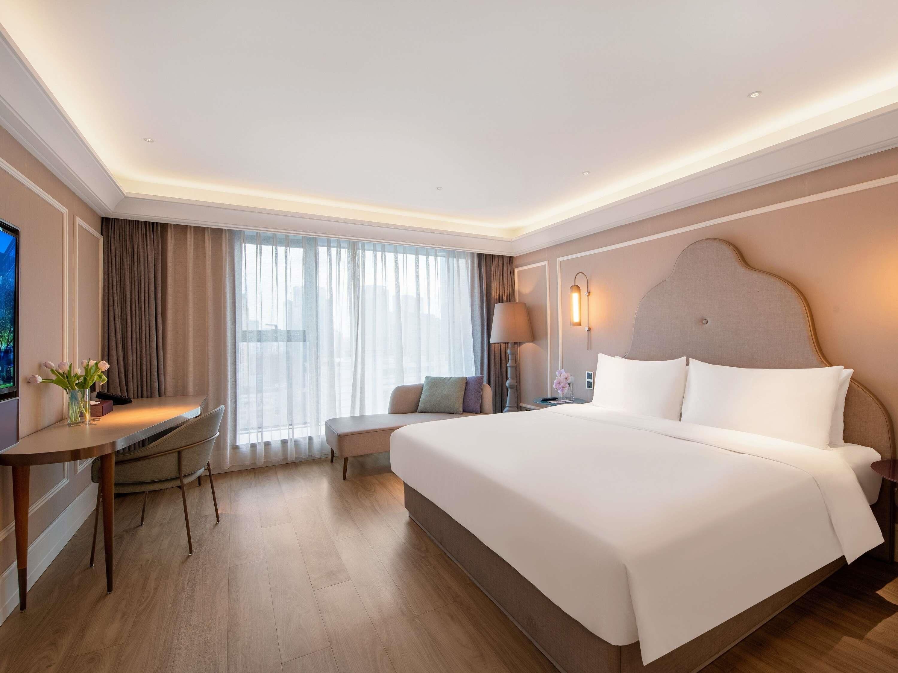 Quarto de hotel com uma cama grande, uma escrivaninha, uma cadeira, um abajur e vista para a cidade pela janela.