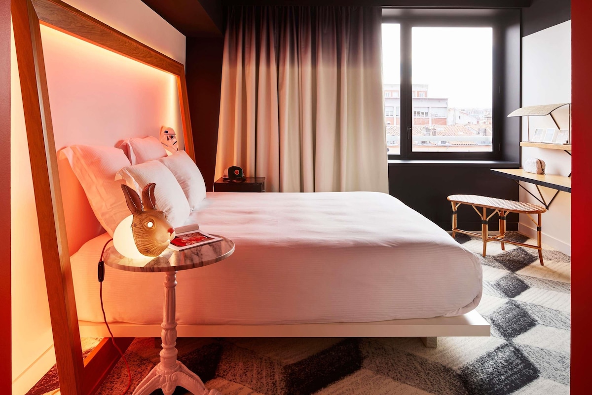Una habitación de hotel moderna con cama, mesita de noche, lámpara y vistas de la ciudad por la ventana.