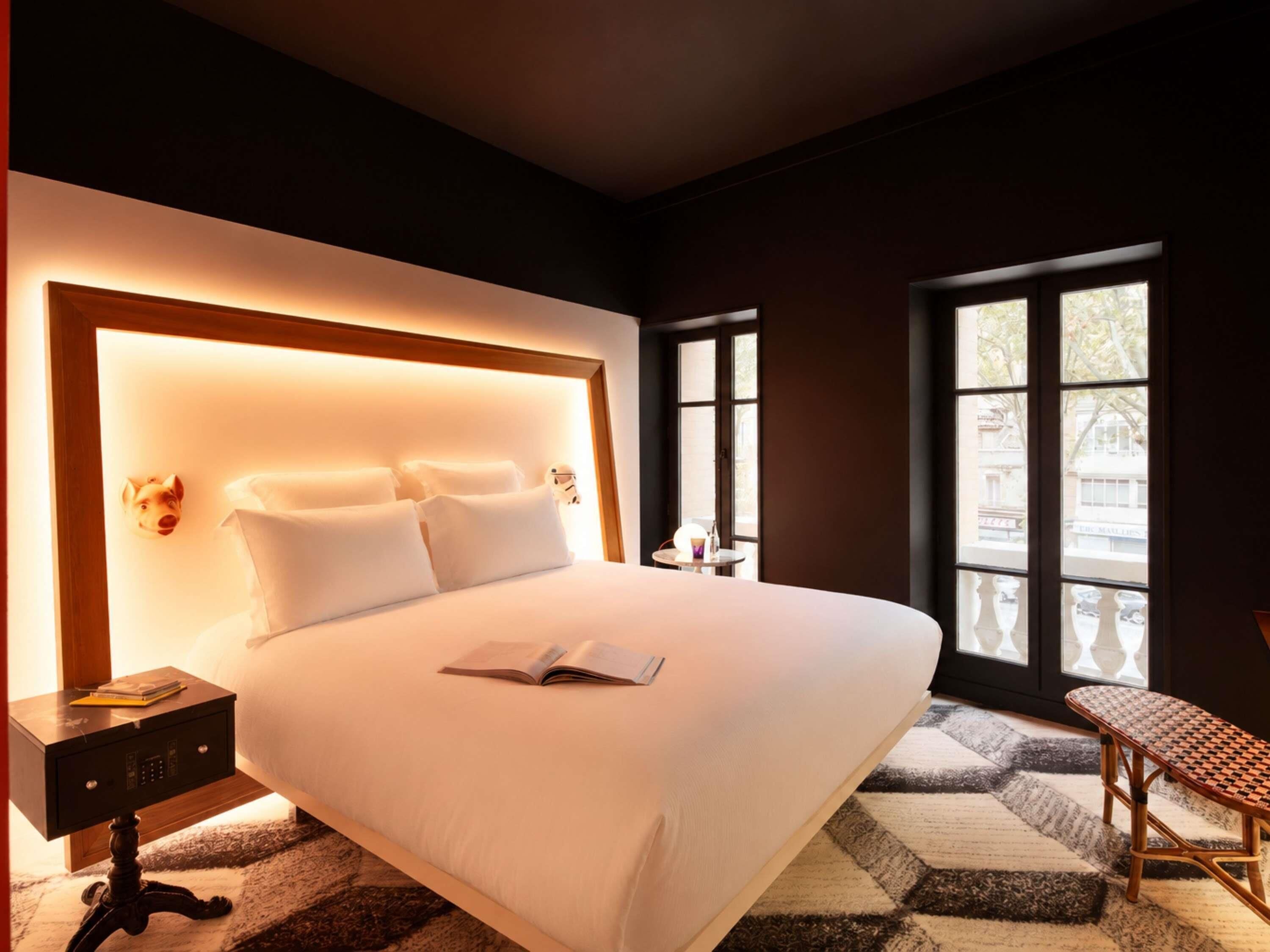 Habitación de hotel con una cama grande, una mesita de noche con lámpara, una silla y una ventana con vistas a edificios.