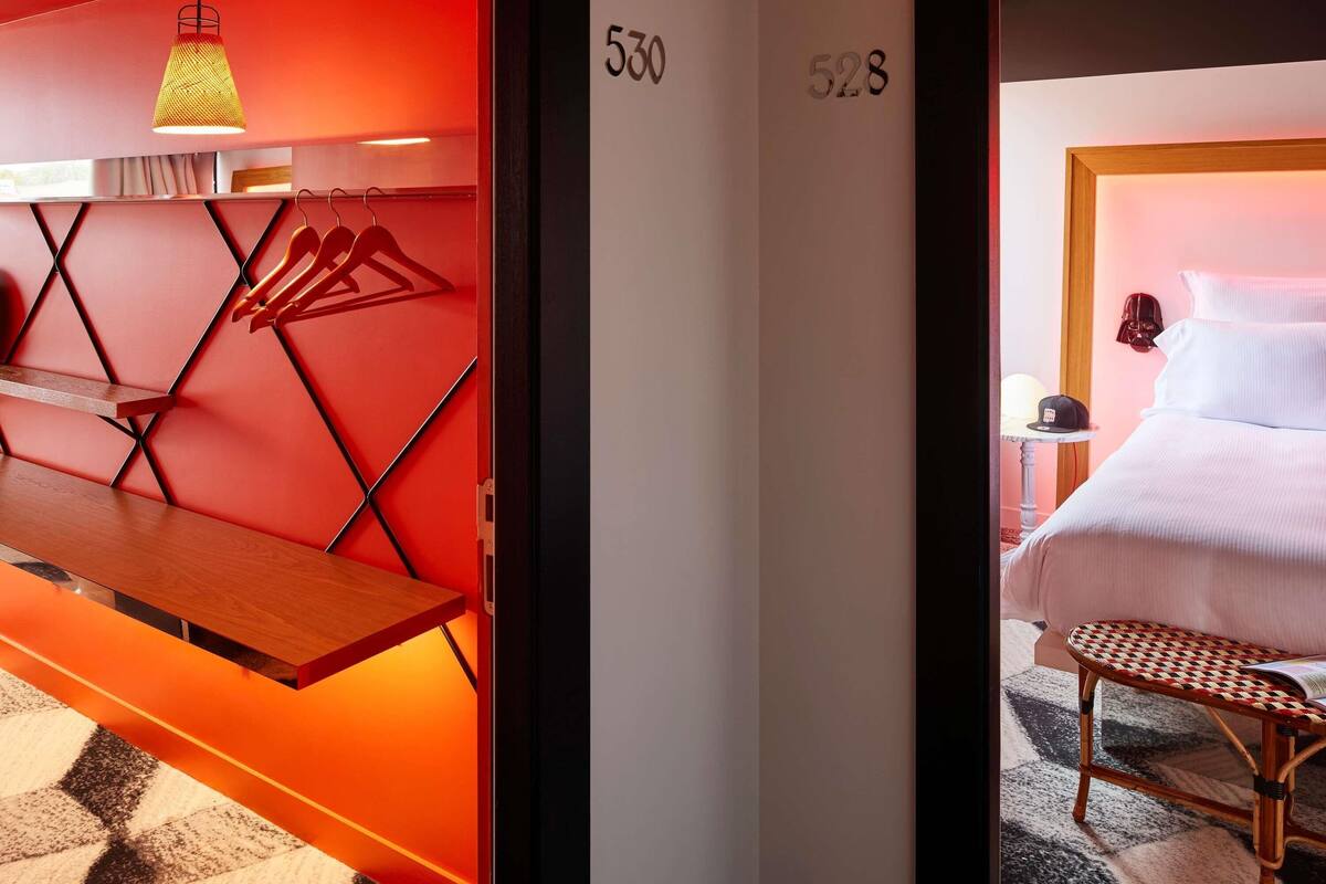 Una habitación de hotel con una pared de acento roja, una cama con ropa blanca, un banco de madera, un perchero en la pared y una lámpara colgante.