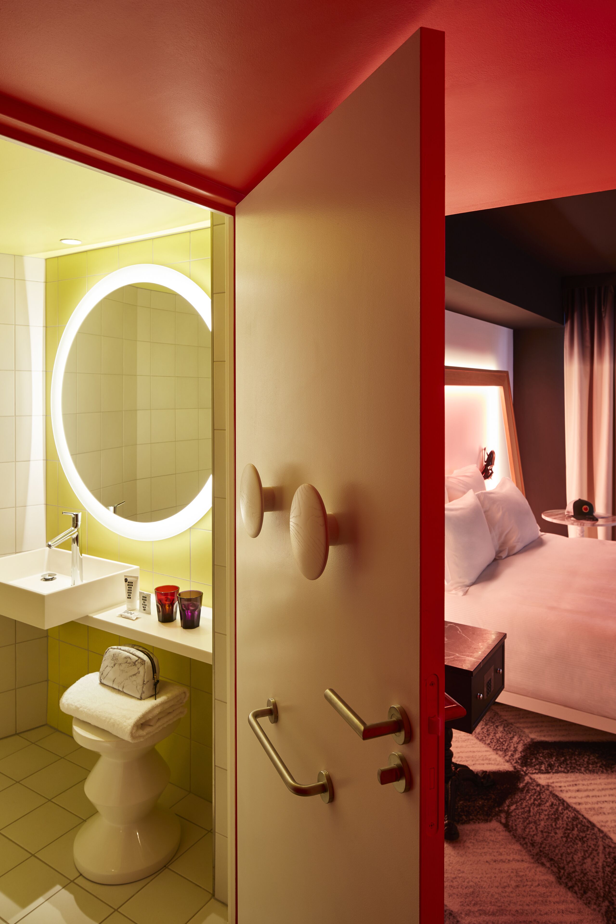 A hotel room with a bathroom featuring a toilet, sink, and mirror with a circular light. The room has a bed with a headboard and a small bedside table.