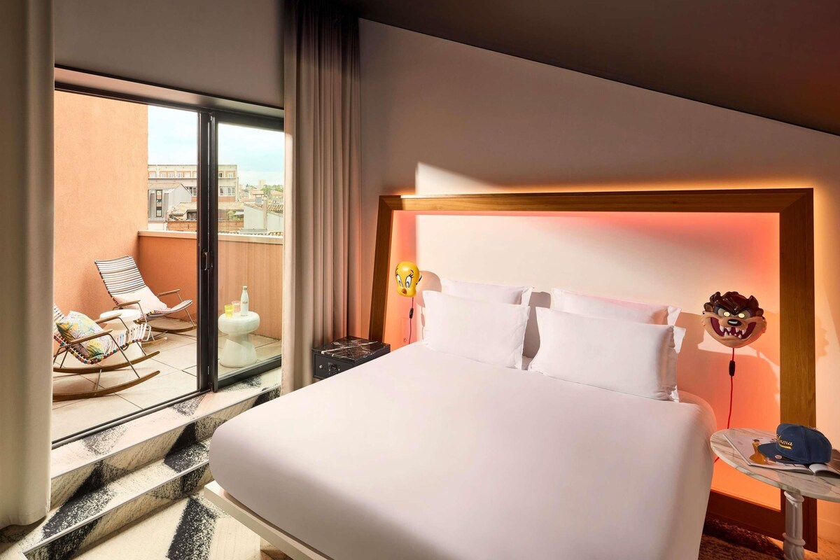 Habitación de hotel con una cama grande, un balcón con sillas y vistas a la ciudad.