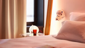 Une chambre d’hôtel avec un lit, une table de chevet sur laquelle se trouvent une bouteille et un verre, une fenêtre avec des rideaux et une tête de lit ornée d’un casque.
