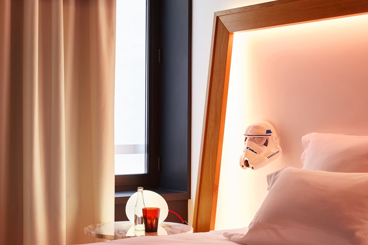 Hotelkamer met een bed, nachtkastje met fles en glas, raam met gordijnen en een hoofdeinde met helm.
