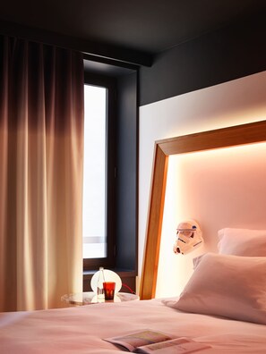 Hotelkamer met een bed, nachtkastje met fles en glas, raam met gordijnen en een hoofdeinde met helm.
