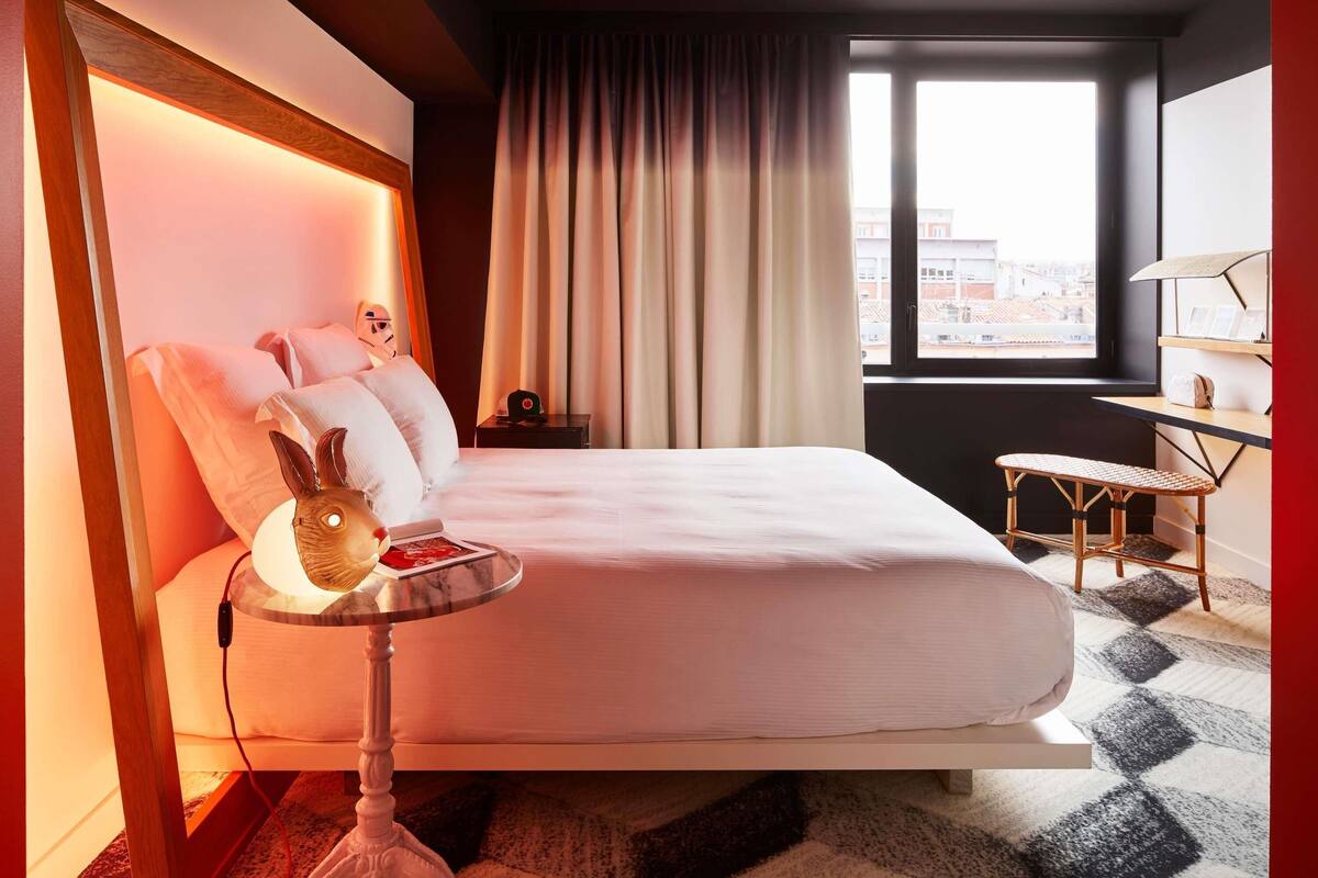 Una habitación de hotel moderna con cama, mesita de noche, lámpara y vistas de la ciudad por la ventana.