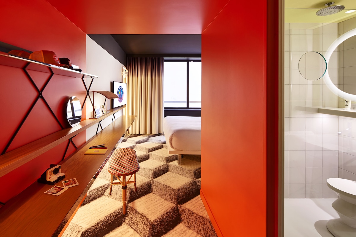 Een moderne hotelkamer met een geruit tapijt, een bed, een bureau met een stoel, en een groot raam.