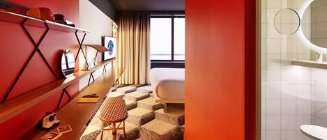 A modern hotel room with a checkered carpet, a bed, a desk with a chair, and a large window.