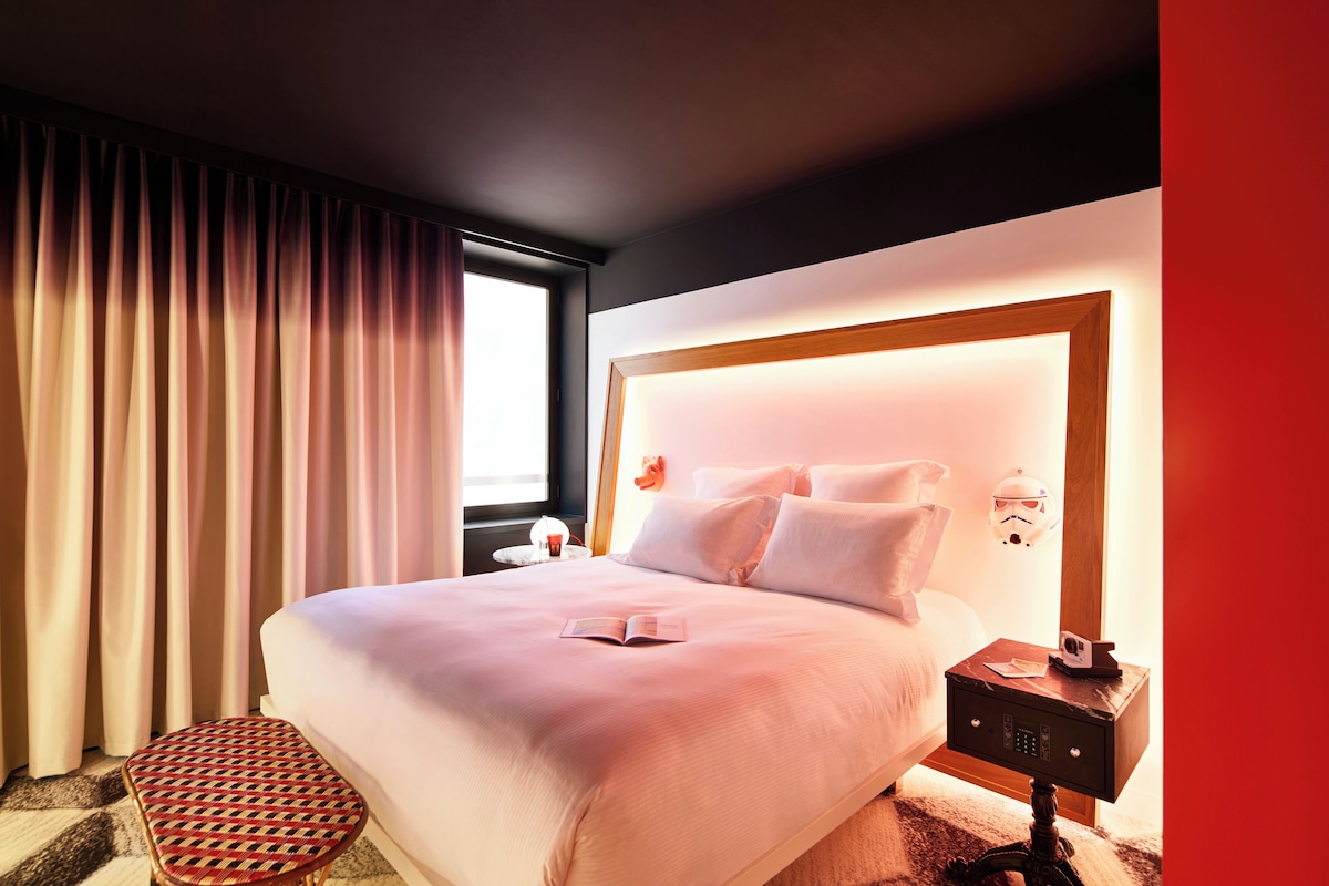 Een hotelkamer met een groot bed, een nachtkastje, een klein krukje en een raam met gordijnen.
