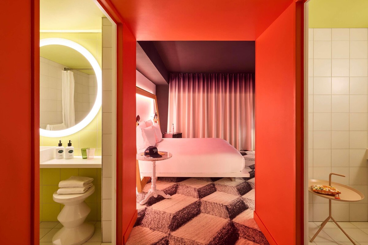 Una habitación de hotel moderna con cama, baño con espejo redondo y una pequeña mesa con una pizza.